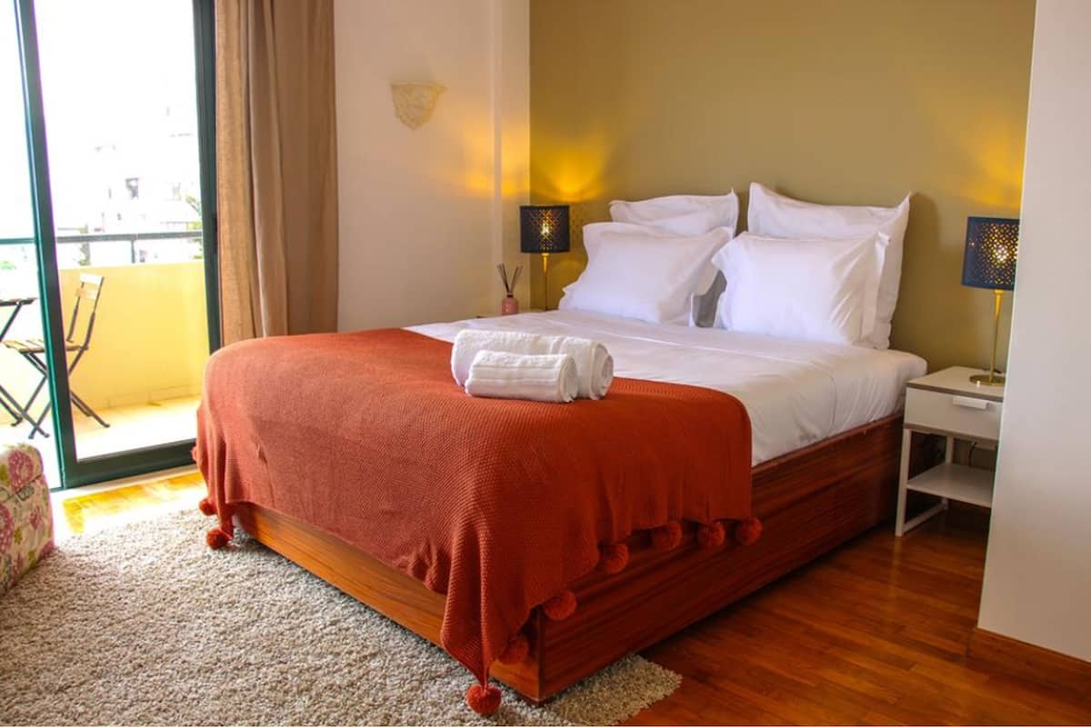 A neatly made bed with white linens and a red blanket, a small bedside table with a lamp, and a view of the outdoors through a sliding door.