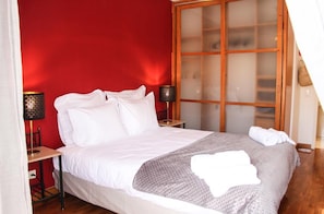 A neatly made bed with white linens, a gray blanket, and a rolled towel on the side. There are two bedside tables with lamps, and a large window with frosted glass panels. - Alameda Garden (Faro)