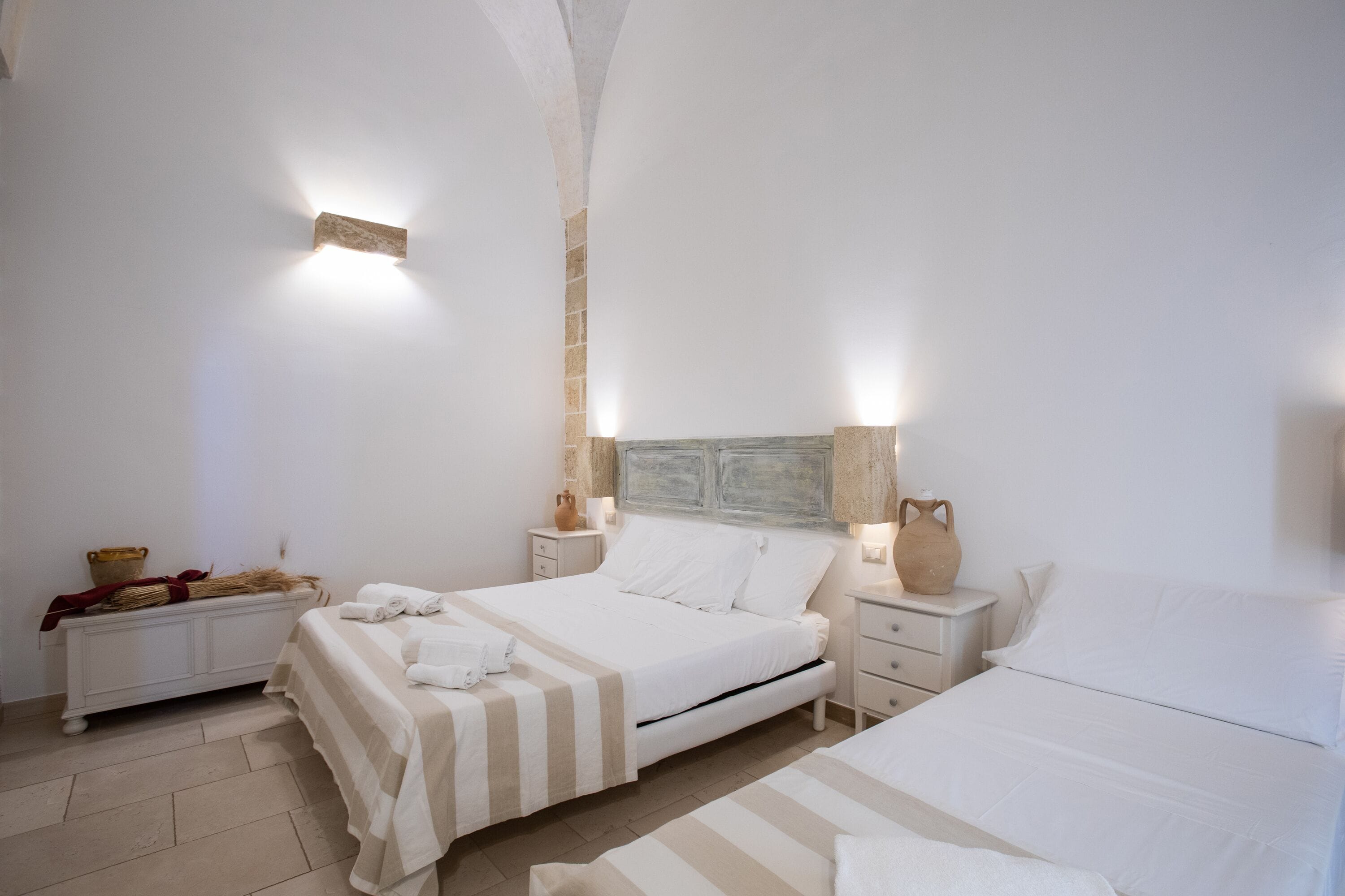 A bedroom with two beds, white linens, a nightstand with a vase, and a wall-mounted light fixture.