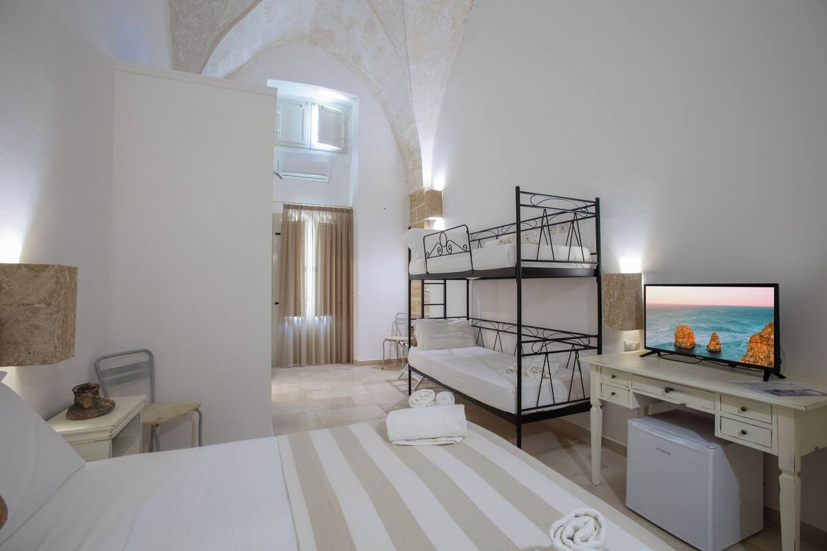 A hotel room with a bunk bed, a desk with a computer, a television, and a small refrigerator.