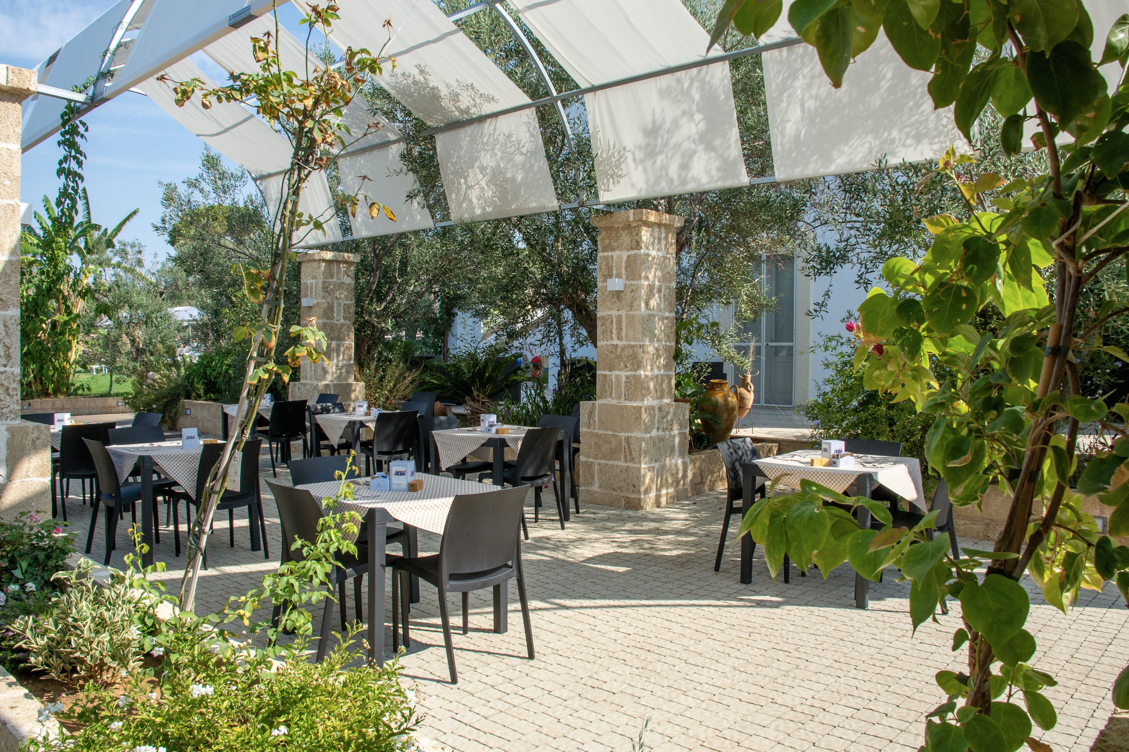 An outdoor patio area with tables and chairs, covered by a translucent canopy, surrounded by greenery and stone pillars.