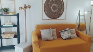 A living room with a mustard-colored sofa, two patterned pillows, a woven wall hanging, a wooden ladder, and a shelf with books and a plant. - Imperia, two-bedroom apartment, nature, and reax (Imperia)