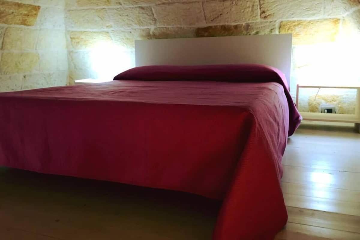 A bedroom with a stone wall, a wooden floor, and a bed with a red bedspread.