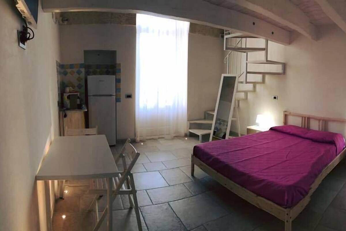 A bedroom with a bed, a desk, a chair, a small table, a nightstand, a lamp, and a staircase.