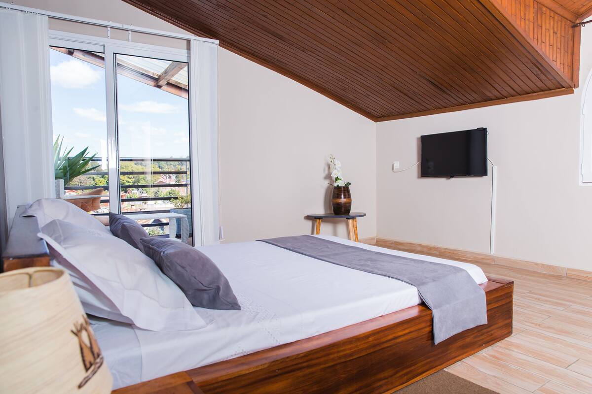 A bedroom with a wooden bed, a TV on the wall, and a view of the outdoors.