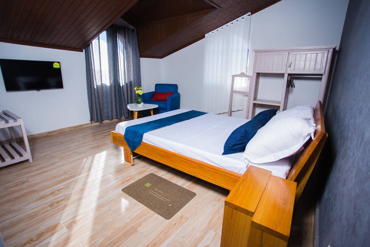 A bedroom with a wooden bed, a chair, a small table, and a TV mounted on the wall.