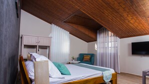 Ein Schlafzimmer mit Holzboden, einem Bett mit weißer und grüner Bettdecke, einem Nachttisch aus Holz, einem kleinen Tisch mit einer Vase, einem türkisfarbenen Sessel und einem Fernseher an der Wand.