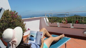 A person lying on a sun lounger reading a book, with a view of the sea and hills in the background.