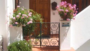 A white building with a wooden door and shuttered windows. There are potted plants with pink flowers in front of the door.