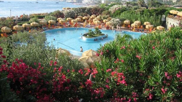 A pool surrounded by umbrellas and greenery, with a view of the sea and buildings in the background.