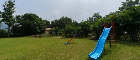 A playground with a blue slide and a red seesaw in a grassy area with trees.