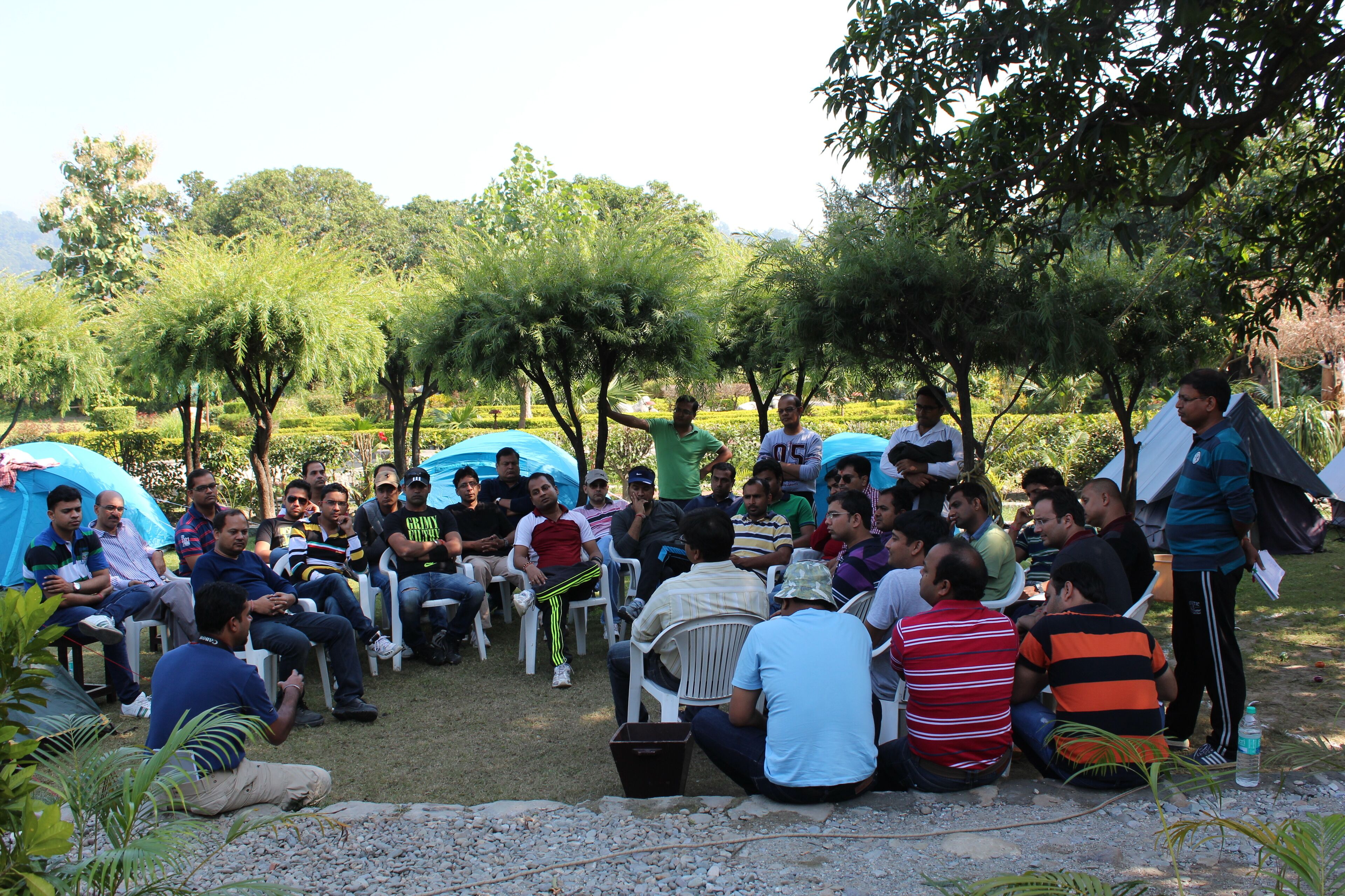 A group of people seated outdoors in a casual setting with tents and trees in the background.