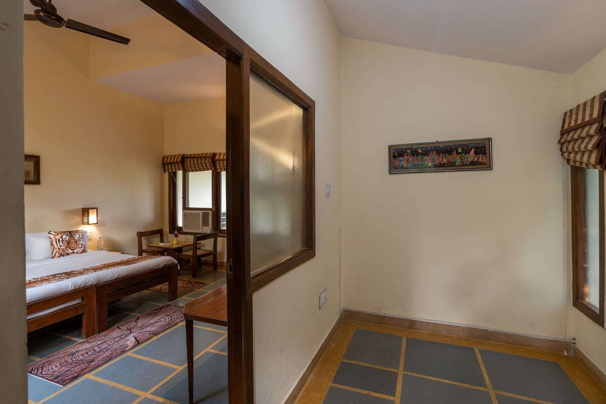 Una habitación con cama, escritorio y silla. Hay una ventana con cortinas, un cuadro en la pared y una puerta que da a otra habitación.