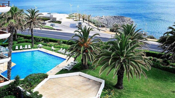 A pool with lounge chairs, palm trees, and a view of the sea. - Ocean View-São Pedro Estoril - Checkinhome (São Pedro do Estoril)