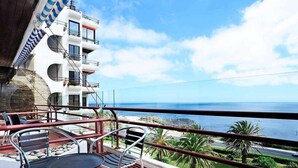 A balcony with a view of the ocean, featuring a table and chairs, and a building with multiple balconies in the background. - Ocean View-São Pedro Estoril - Checkinhome (São Pedro do Estoril)