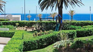 A landscaped garden with a palm tree, benches, and a playground. - Ocean View-São Pedro Estoril - Checkinhome (São Pedro do Estoril)