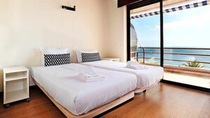 A double bed with white linens and black patterned pillows, a small bedside table, and a view of the ocean through large windows. - Ocean View-São Pedro Estoril - Checkinhome (São Pedro do Estoril)