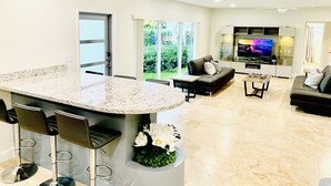 A modern kitchen with a large island, bar stools, a sofa, and a TV in a living area with a view of greenery. - SHORT WALK TO BEACH AND RESTAURANTS HEATED POOL (Lauderdale-By-The-Sea)
