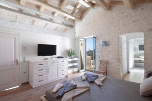 Un dormitorio con techo de madera, un televisor sobre un tocador blanco, una cama con mantas enrolladas y vistas al exterior a través de una puerta de cristal.
