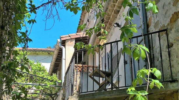 Una escalera de piedra que lleva a un edificio con balcón, rodeado de vegetación y un cielo azul despejado.