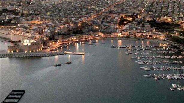 A marina with numerous boats docked, a waterfront promenade, and buildings along the shore.