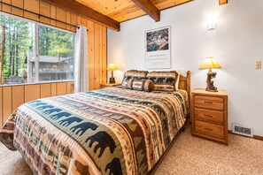 A bedroom with a large bed, a wooden dresser, two bedside lamps, a window with a view of trees, and a framed picture on the wall.