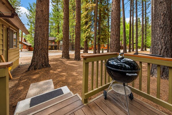 Una terraza de madera con una parrilla, rodeada de árboles altos y un edificio residencial.