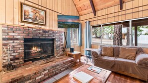 A cozy living room with a brick fireplace, a sofa, a coffee table, and a painting above the fireplace. - Bear Crossing - Walk to Slopes, Foosball Table, Barbecue (Big Bear Lake)
