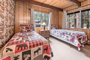 Un dormitorio de estilo cabaña con dos camas, cada una cubierta con un edredón estampado con motivos animales, una mesita de noche, una lámpara y ventanas grandes que ofrecen vistas del bosque circundante.