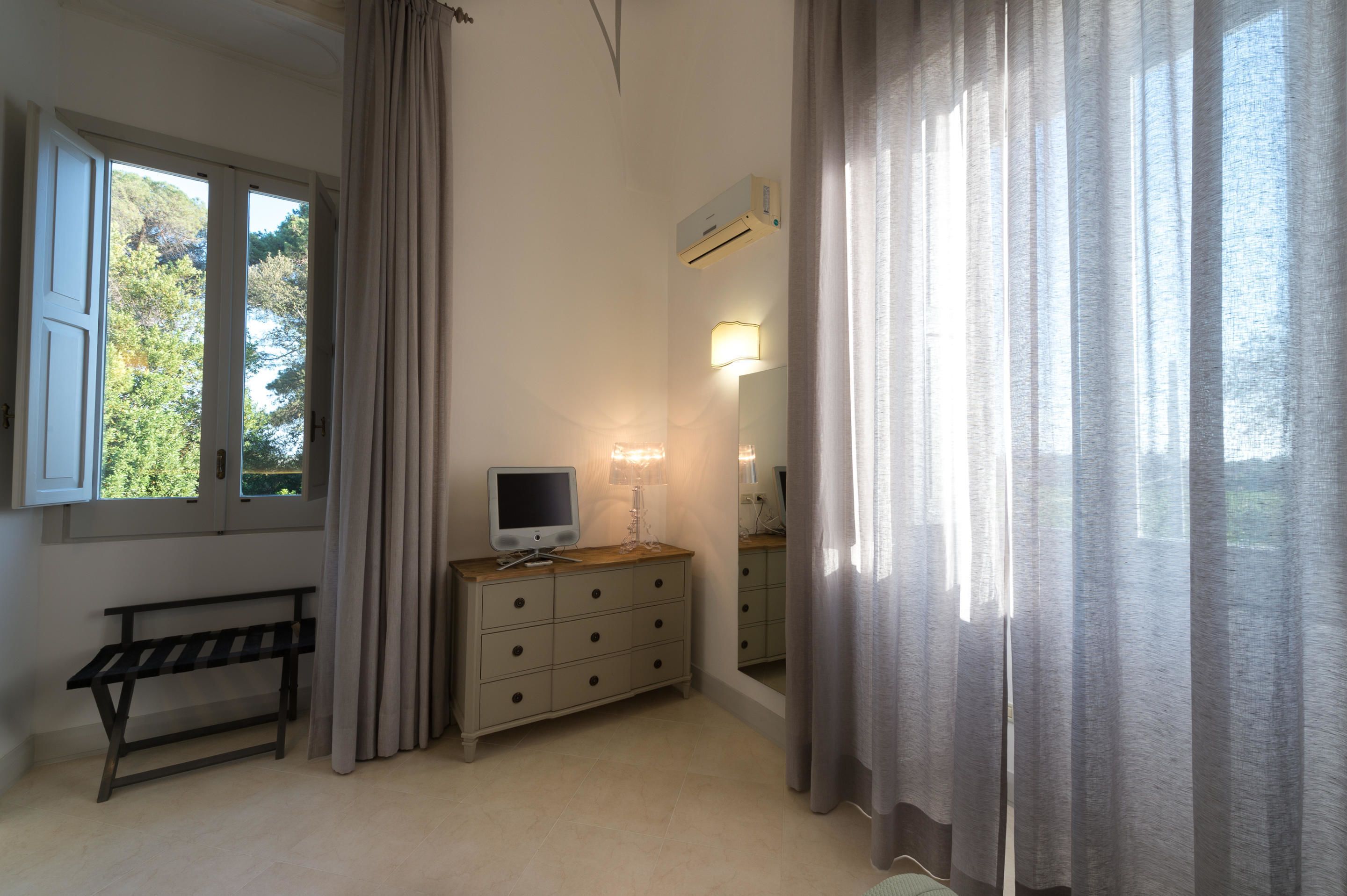 A bedroom with a bed, a bedside table, a lamp, a TV, and a window with curtains.