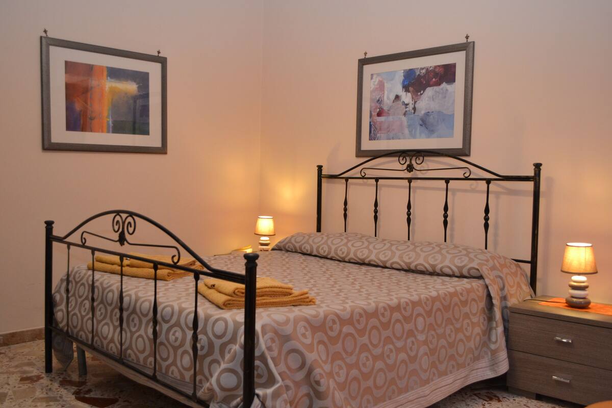 Una cama bien hecha con ropa de cama estampada, una mesita de noche con lámpara y dos cuadros enmarcados en la pared.