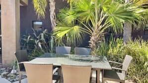 Een buitenterras met rieten meubels en een glazen tafel, omgeven door tropische planten en een stenen gebouw.