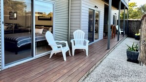Een modern huis met een overdekt terras, schuifdeuren die leiden naar een slaapkamer, en een klein zitgedeelte buiten met twee witte stoelen.
