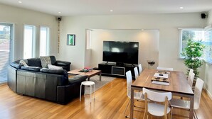 Een moderne woonkamer met een grote flatscreen-televisie, een zwarte leren bank, een houten eettafel met stoelen en een klein, wit krukje.