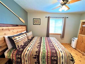 A bedroom with a wooden headboard, a bed with patterned bedding, a ceiling fan, a window with curtains, and a white air conditioning unit. - #2 Trout Cabin at Ponderosa Place (Custer)