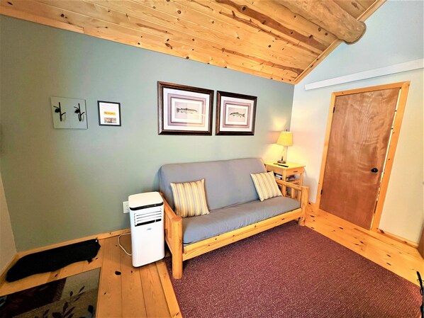 A cozy room with a wooden ceiling, a grey sofa with striped pillows, a small table with a lamp, and framed pictures on the wall. - #2 Trout Cabin at Ponderosa Place (Custer)