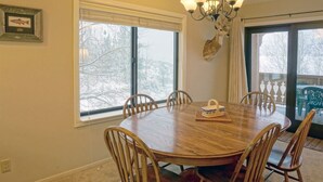A dining room with a round wooden table, six chairs, a chandelier, and large windows with a view of trees. - June Lake Interlaken 12 Cozy, Spacious (June Lake)