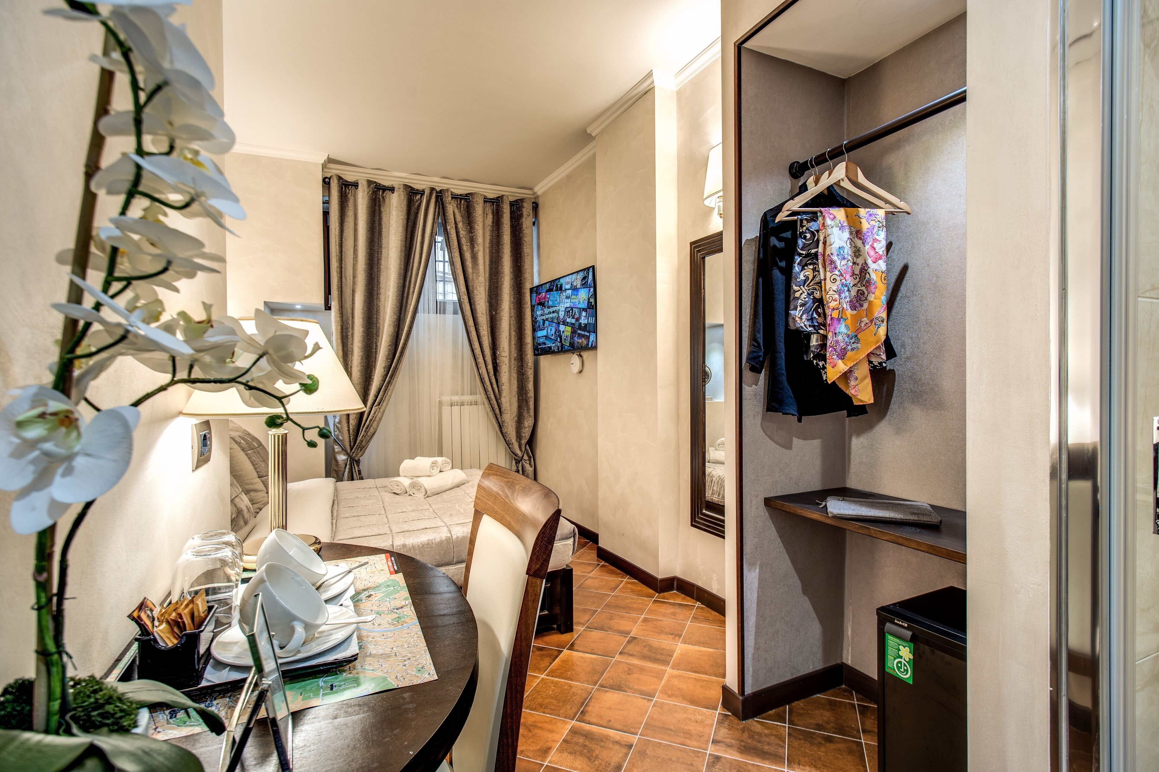 Camera d'albergo con zona pranzo, divano, televisione, specchio, portasciugamani e un piccolo frigorifero.