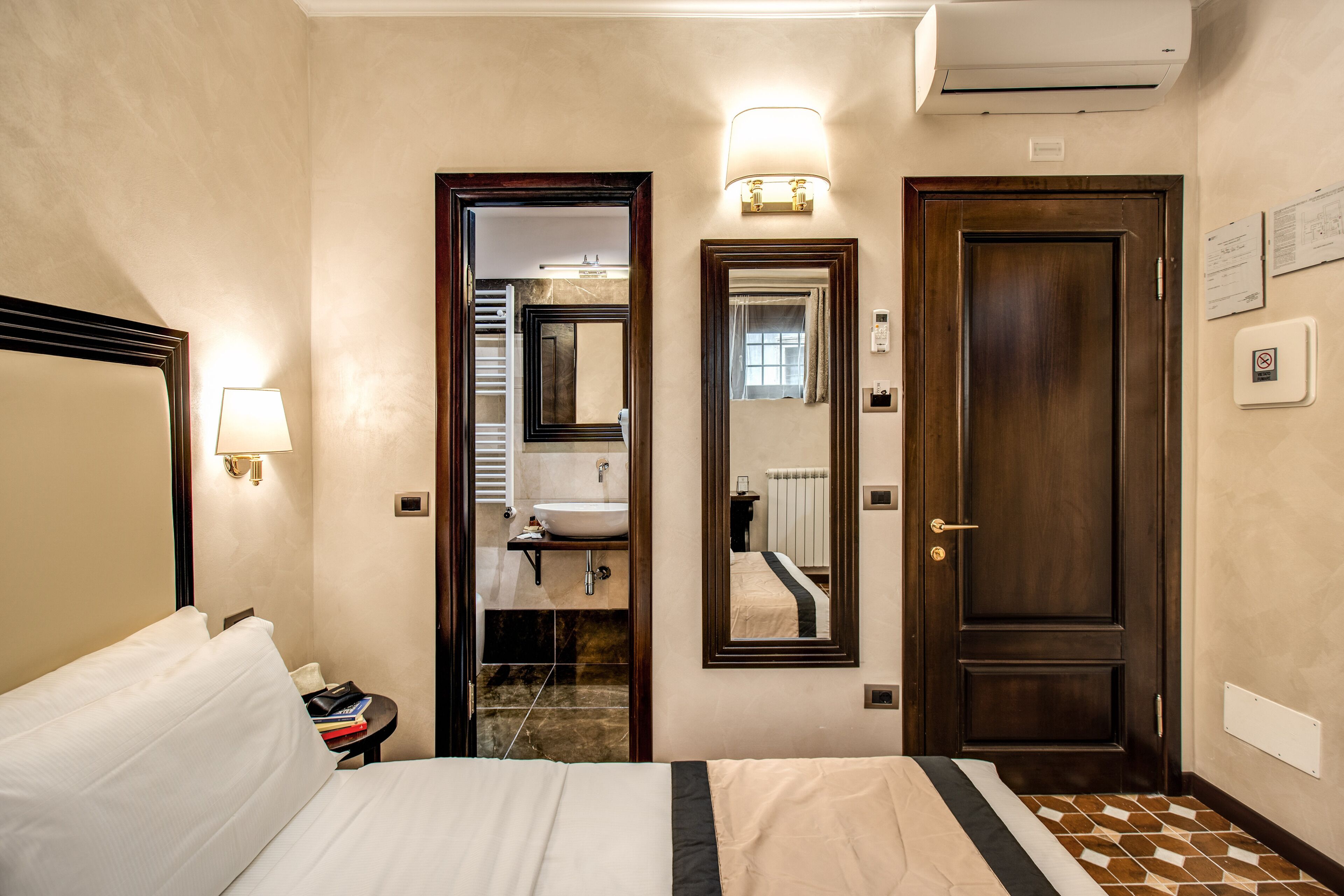 A hotel room with a bed, bedside lamp, a mirror, a door, and a wall-mounted air conditioning unit.