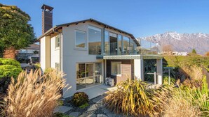 A modern two-story house with large windows, a balcony, and a mountain backdrop.