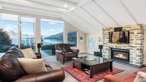 A modern living room with a stone fireplace, leather sofas, and large windows offering a scenic view of mountains and a lake.