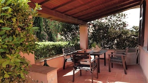 Um pátio externo com mesa e cadeiras de madeira, rodeado por vegetação e piso de azulejos.