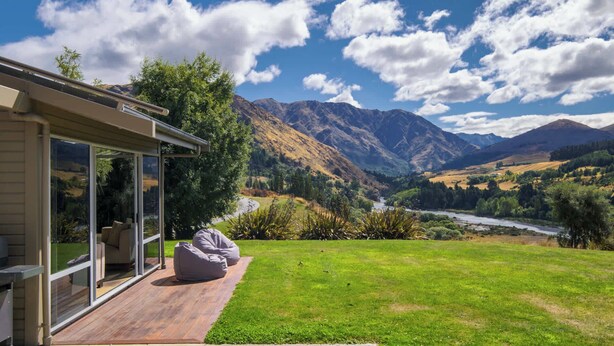 Ein modernes Haus mit Glastür, die auf eine Terrasse führt, mit Blick auf einen Fluss und Berge.