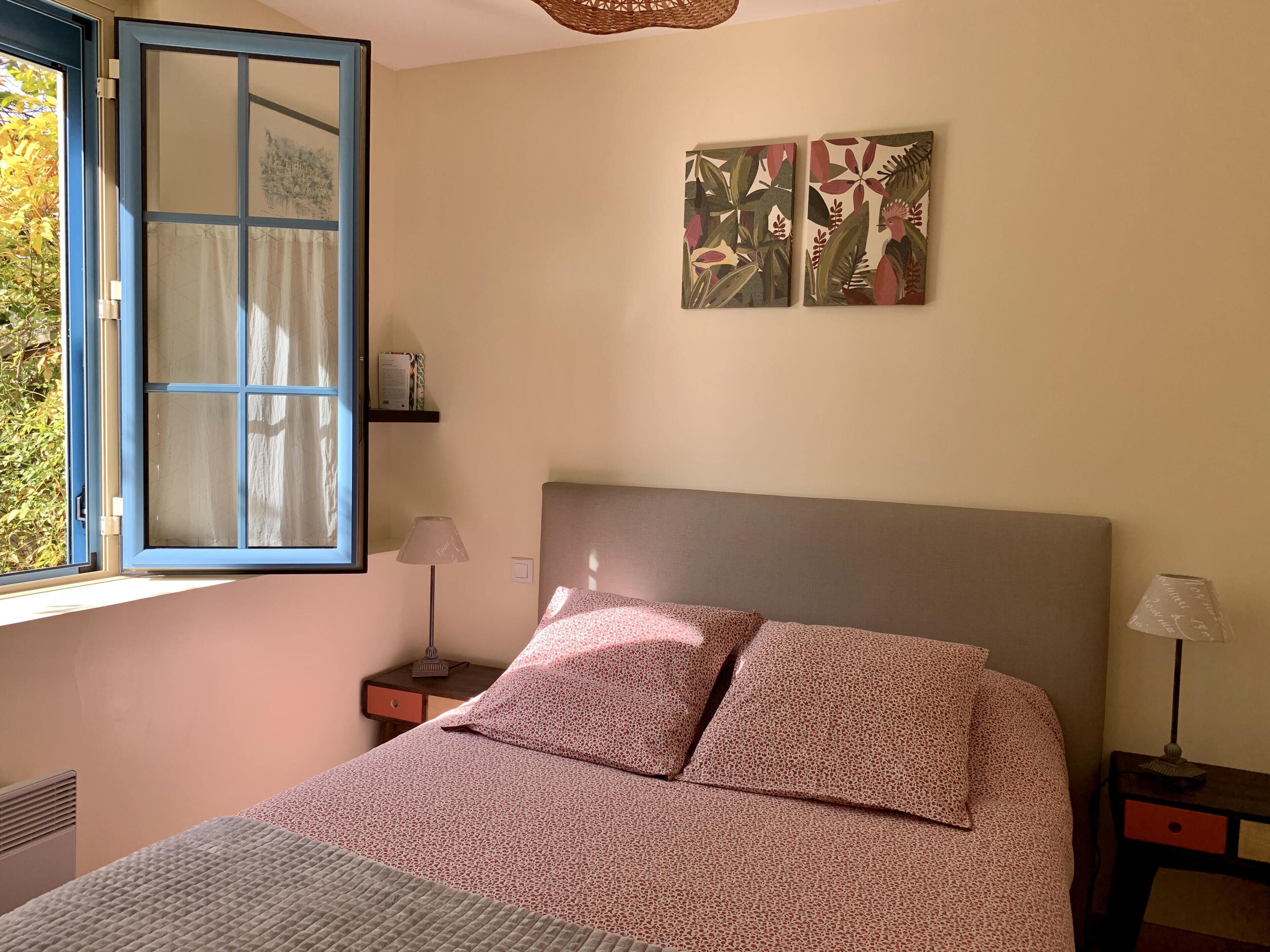 A bedroom with a bed, a window with curtains, a bedside table, a lamp, and wall art.