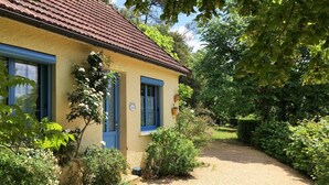 Ein gelbes Haus mit blauen Fensterläden und rotem Dach, umgeben von viel Grün und einem Kiesweg.