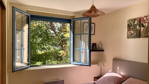 Ein Schlafzimmer mit offenem Fenster, einem Bett mit gemusterten Betttextilien, einem Nachttisch mit Lampe sowie einem Wandregal mit Büchern und einer kleinen Statue.
