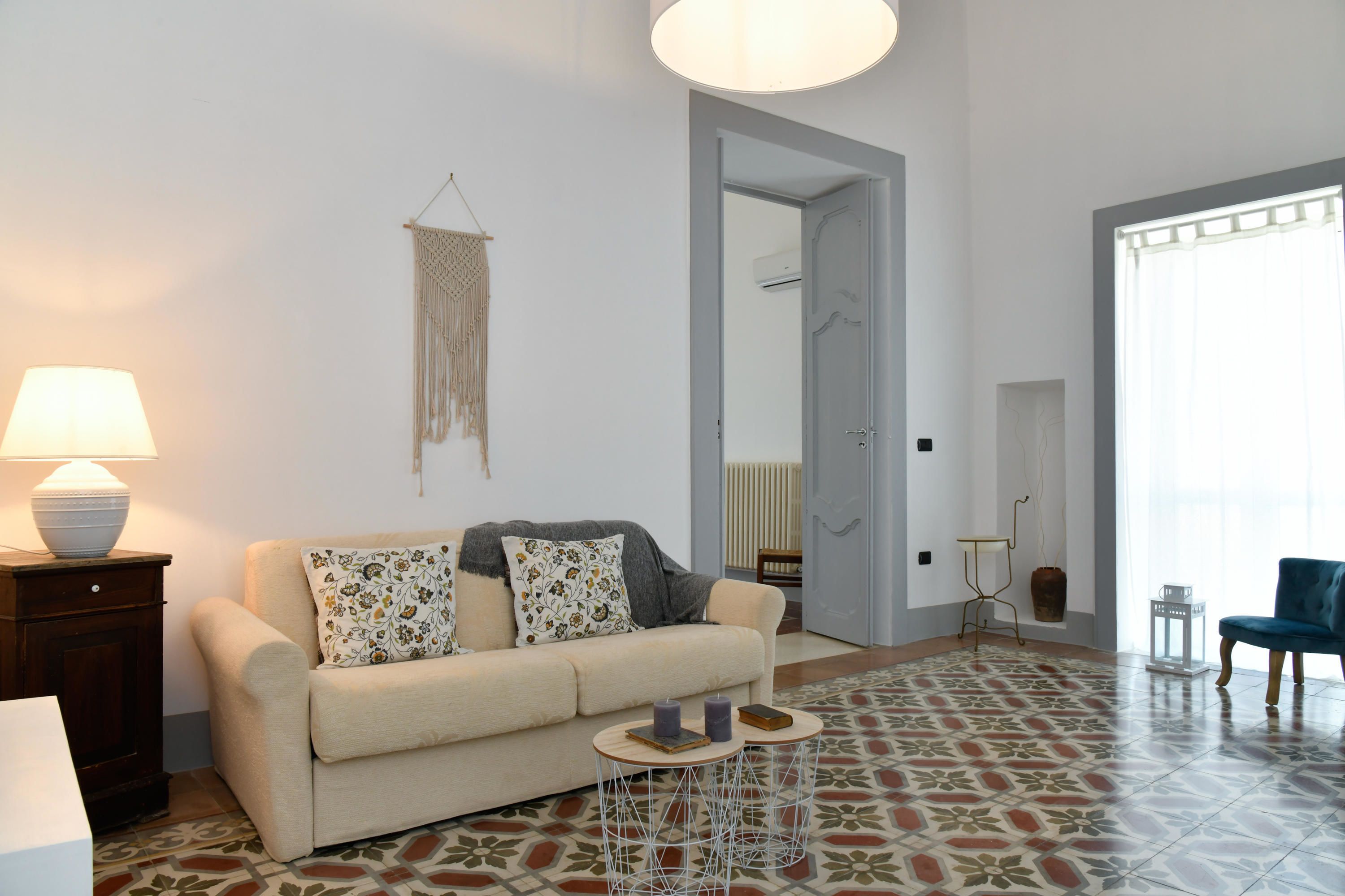 Uma sala de estar com sofá, duas mesinhas laterais, luminária e piso estampado.