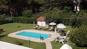 Una piscina con sillas de descanso y sombrillas, rodeada de setos y árboles bien cuidados.