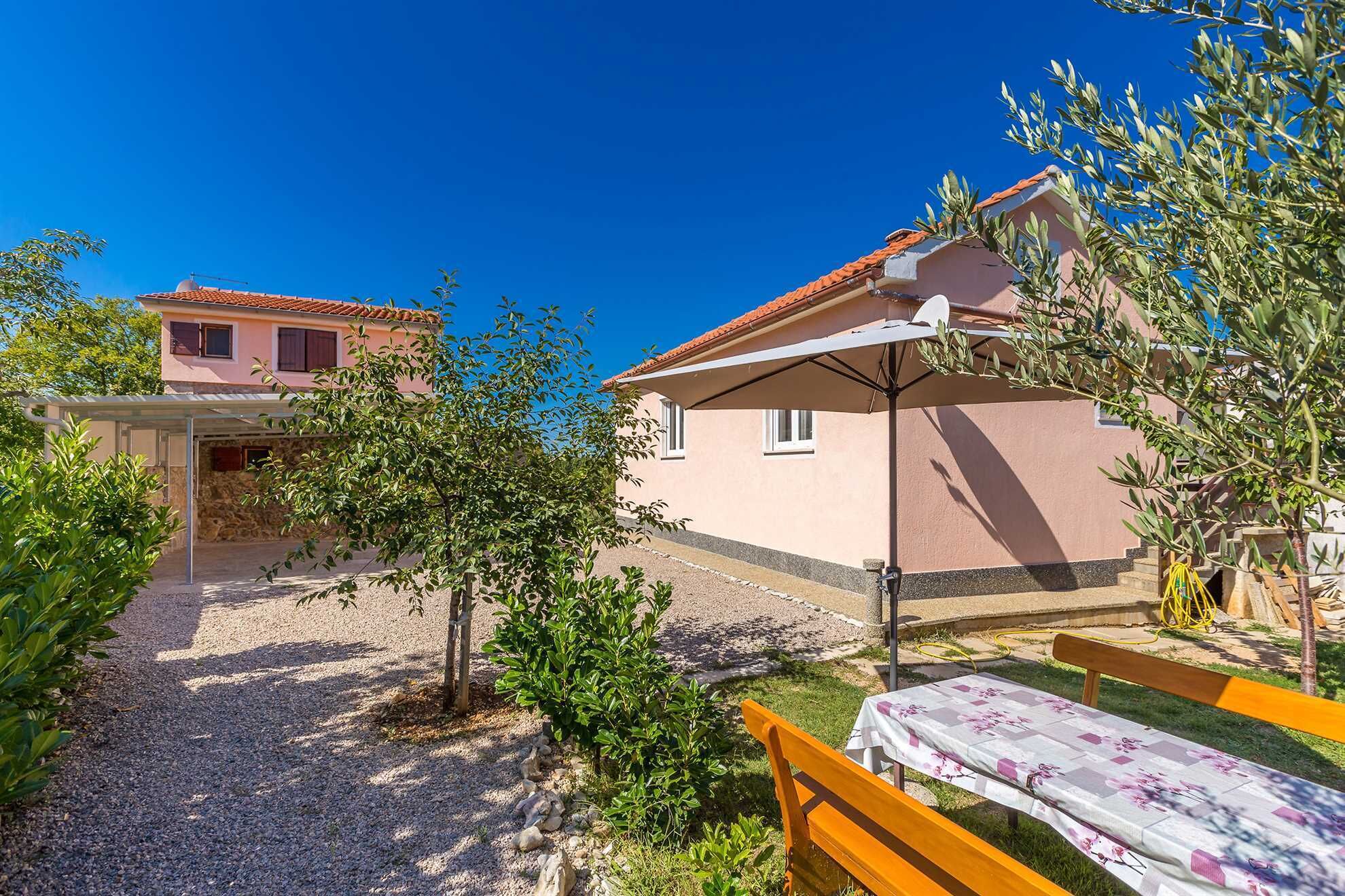 Ein Wohnhaus mit überdachter Terrasse, gepflasterter Auffahrt und kleinem Garten mit Bank und Tisch.