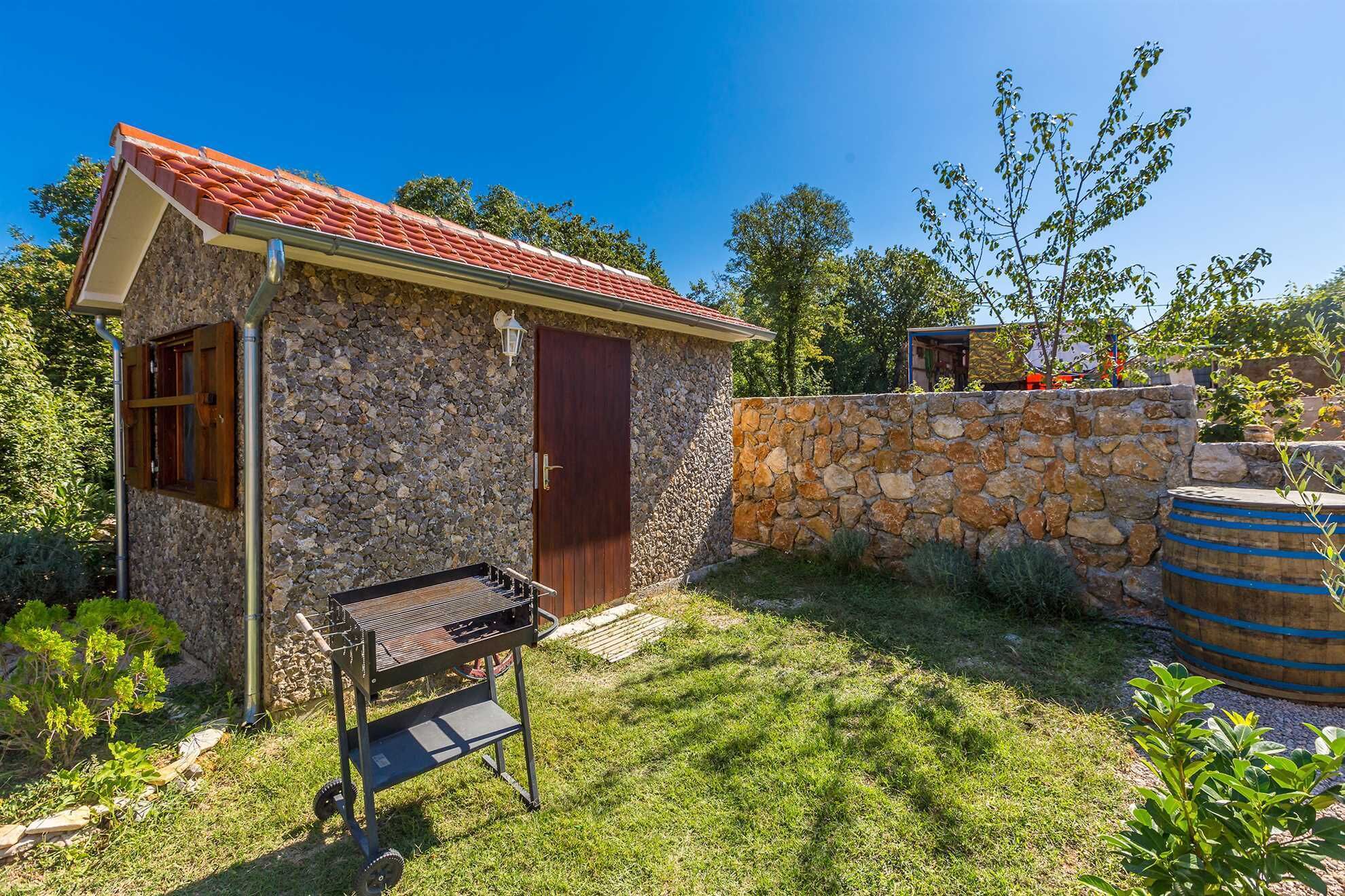 A stone house with a red-tiled roof, a wooden door, and a barbecue grill in the yard.